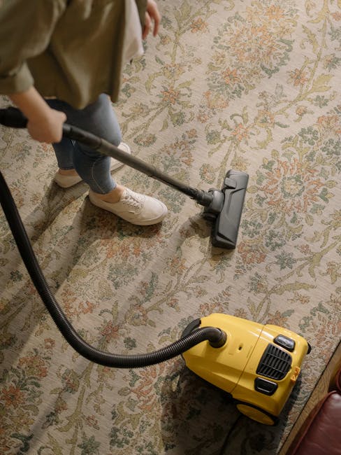 A person using a handheld vacuum cleaner connected to a yellow canister to perform surface cleaning on a traditional patterned carpet in a residential living space. The individual is wearing casual attire and white sneakers, focusing on vacuuming the light-colored, floral design rug. The room is well-lit, with the carpet appearing clean and free of visible dust or debris. The image emphasizes professional deep cleaning techniques employed by Richmond Carpet Cleaning, as seen in the context of their services for domestic and commercial environments on the page titled 'Kew Road carpet cleaning experts serving Richmond TW9' noted on the website richmondcarpetcleaning.org.uk.