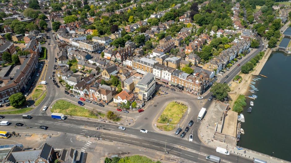 An aerial view of Kew Road in Richmond showing a mix of residential and commercial buildings with brick facades, tiled roofs, and large windows. The street is busy with cars and buses moving along the road, which runs parallel to the river Thames visible on the right side of the image. Adjacent to the river is a promenade with boats docked along the water, and green spaces with trees lining the area. The lighting is bright, suggesting a sunny day with clear skies, highlighting the well-maintained urban environment. Richmond Carpet Cleaning emphasizes maintaining cleanliness and hygiene in such busy residential and commercial spaces, offering surface cleaning and deep sanitisation services to uphold hygiene standards in local properties.