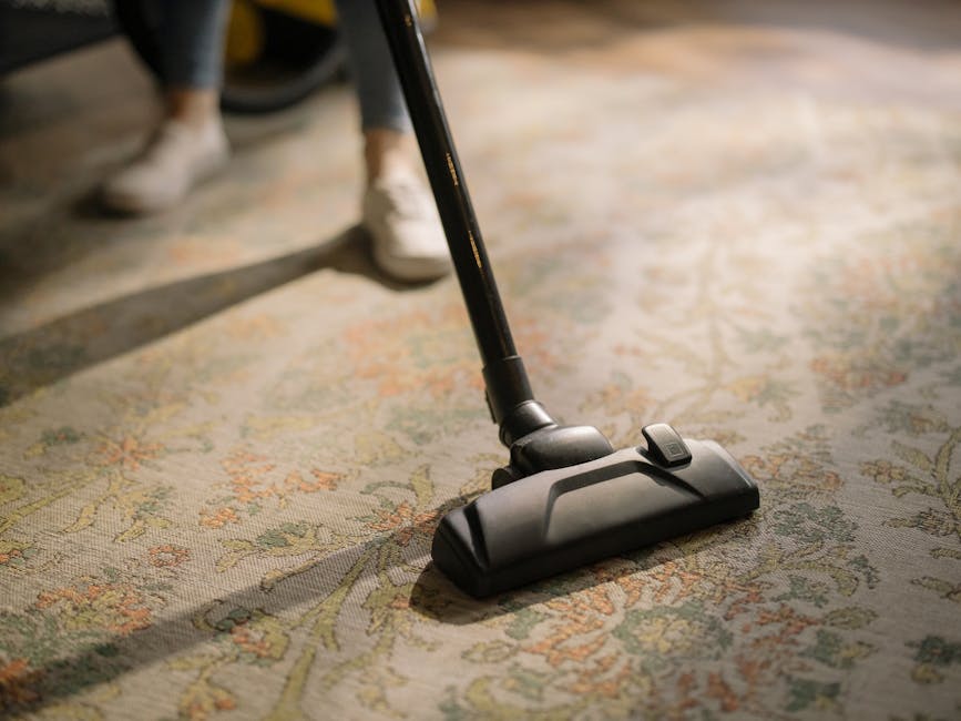 A person using a handheld vacuum cleaner connected to a yellow canister to perform surface cleaning on a traditional patterned carpet in a residential living space. The individual is wearing casual attire and white sneakers, focusing on vacuuming the light-colored, floral design rug. The room is well-lit, with the carpet appearing clean and free of visible dust or debris. The image emphasizes professional deep cleaning techniques employed by Richmond Carpet Cleaning, as seen in the context of their services for domestic and commercial environments on the page titled 'Kew Road carpet cleaning experts serving Richmond TW9' noted on the website richmondcarpetcleaning.org.uk.