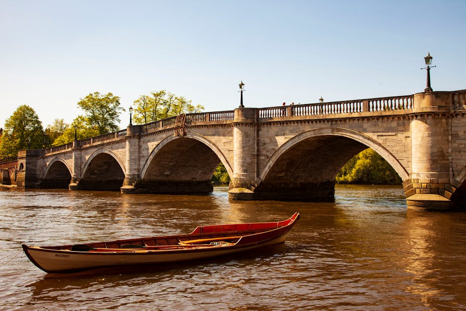 A stone bridge with multiple arches spans across a river in an urban park, featuring ornate street lamps along the balustrade. The river water is calm, with slight ripples, and a red and white boat is moored in the foreground. The surface of the bridge appears cleaned and well-maintained, with sunlight illuminating the stonework and casting gentle reflections on the water. Surrounding trees with green foliage are visible in the background, indicating a lush environment. The scene exudes a sense of cleanliness and order typical of a well-kept public space, consistent with deep cleaning and surface maintenance practices that firms like Richmond Carpet Cleaning promote for hygiene and appearance, even though the image portrays an outdoor setting.