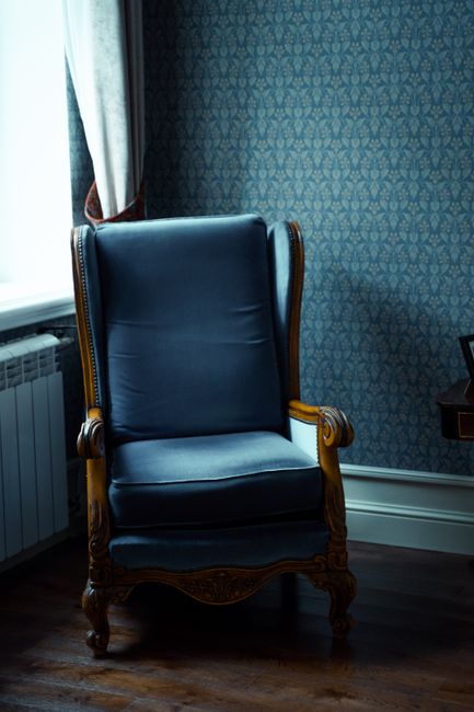 An elegant wooden armchair with a dark blue leather cushion, positioned beside a window with a light patterned curtain in a room with intricate blue wallpaper. The dark wooden floor is visible beneath the chair, and a radiator is partially seen beneath the window. The setting suggests a well-maintained, period-style interior with a focus on furniture cleaning and surface maintenance, as part of domestic cleaning services offered by Richmond Carpet Cleaning in Richmond. The room appears bright and tidy, emphasizing the importance of regular sanitisation and deep cleaning in historic homes.