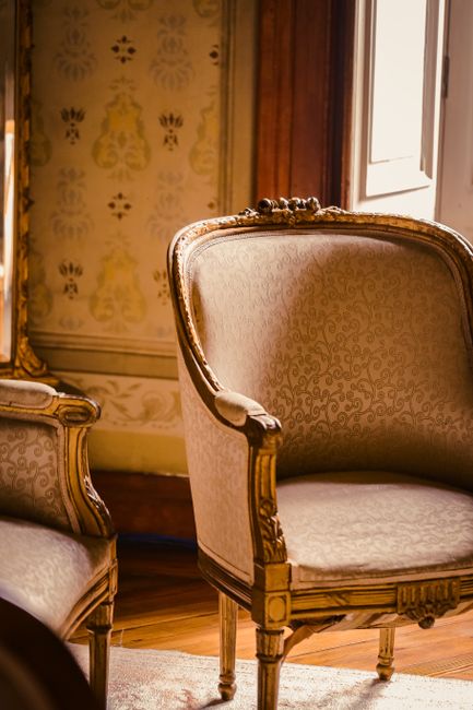 A close-up view of an ornate, vintage armchair with a carved wooden frame and beige upholstered fabric featuring a subtle pattern, positioned in a richly decorated room with patterned wallpaper in shades of gold and cream. Sunlight streams in through a partially visible window, illuminating the chair and highlighting the polished wooden floor beneath. The setting suggests a period home interior, reflecting traditional furnishings and elegant decor, with evidence of professional deep cleaning, such as the absence of dust or stains on the upholstery. Richmond Carpet Cleaning ensures the maintained cleanliness and preservation of such furnishings in line with their domestic cleaning and surface sanitisation services.