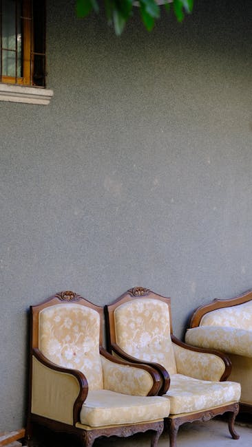 An elegant wooden armchair with a dark blue leather cushion, positioned beside a window with a light patterned curtain in a room with intricate blue wallpaper. The dark wooden floor is visible beneath the chair, and a radiator is partially seen beneath the window. The setting suggests a well-maintained, period-style interior with a focus on furniture cleaning and surface maintenance, as part of domestic cleaning services offered by Richmond Carpet Cleaning in Richmond. The room appears bright and tidy, emphasizing the importance of regular sanitisation and deep cleaning in historic homes.